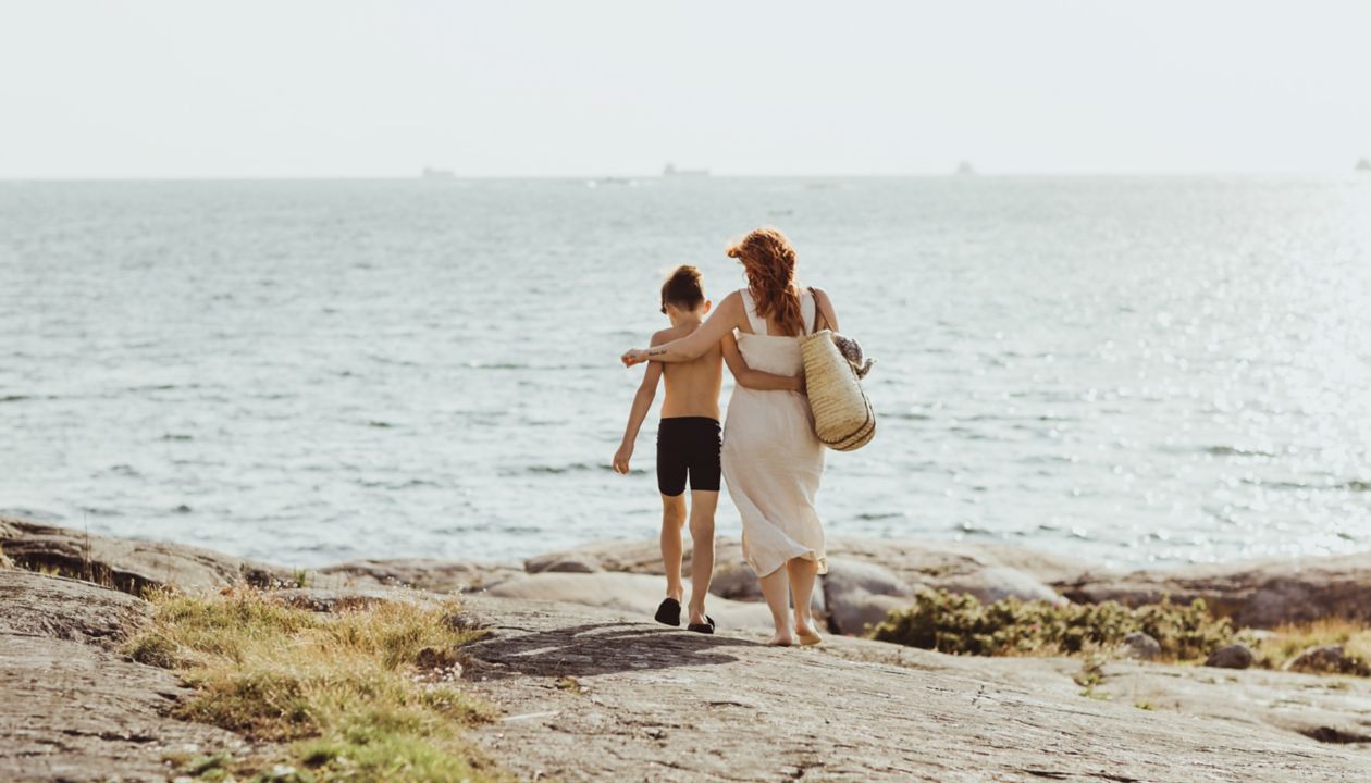 Kvinde med søn set bagfra. De går langs havet på en solskinsdag