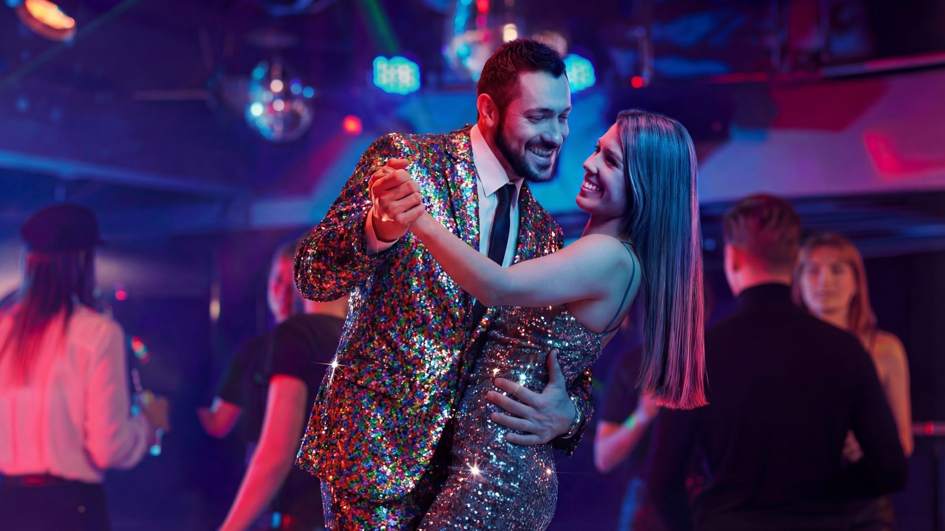 A couple dressed in glittering, sequined outfits is seen dancing closely in a vibrant nightclub setting. The man wears a sparkling suit, while the woman dons a shimmering silver dress. The background features colorful lights, disco balls, and other people enjoying the lively atmosphere.