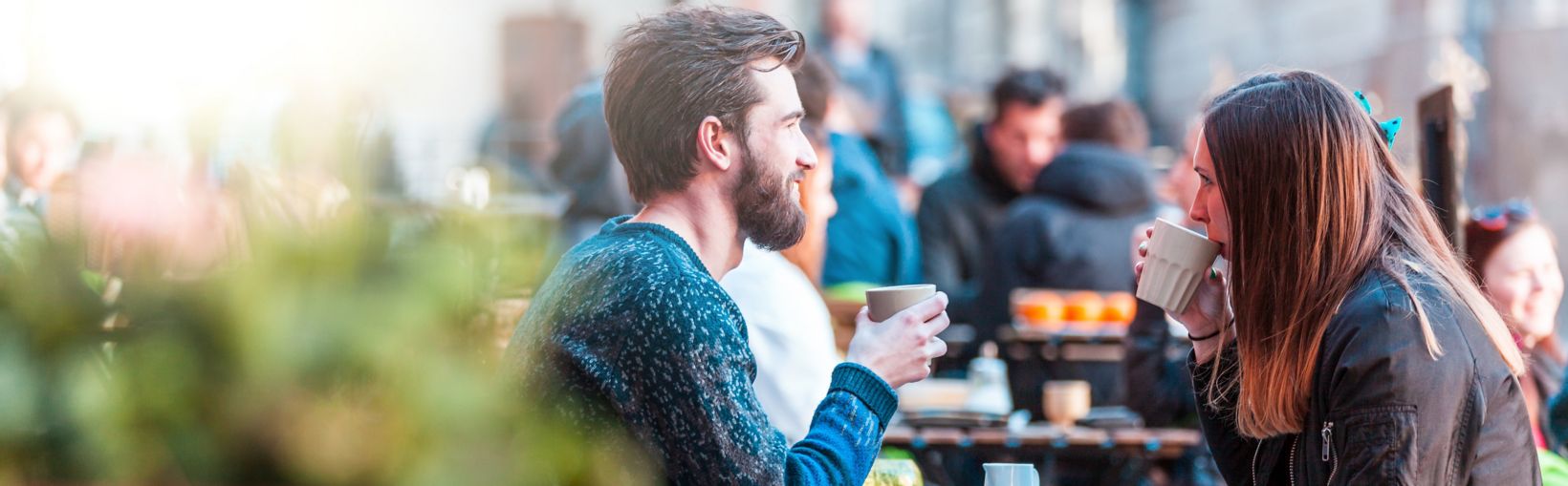 Ung, voksen kvinde med langt, brunt hår og mandlig ven drikker kaffe ved et bord uden for en café på en klar, men kold dag