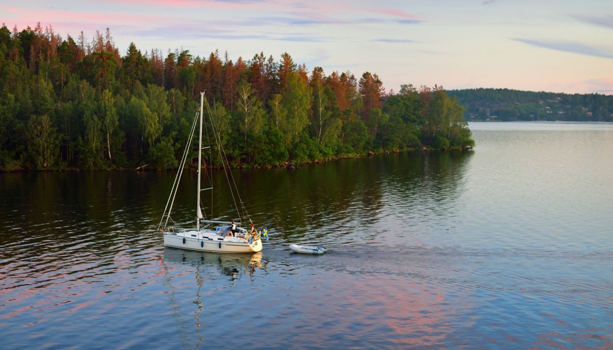 Sloop rigged yacht with inflatable boat sailing at sunset. Panoramic aerial view of the forest lakeshore. Moonrise, glowing clouds. MÃ¤laren lake, Sweden. Summer vacations, nature, sailing, cruise