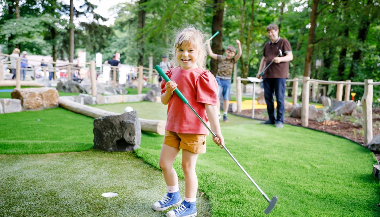 Jauka pirmsskolas meitene, kas spēlē mini golfu ar ģimeni. Laimīgs mazulis, kas izklaidējas ar āra aktivitātēm. Vasaras sports bērniem un pieaugušajiem, ārā. Ģimenes brīvdienas vai kūrorts