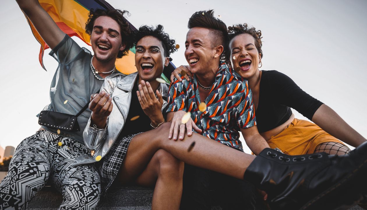 Young people celebrating pride while sitting together. Four members of the LGBTQ+ community smiling cheerfully while raising the pride flag. Group of queer individuals celebrating together outdoors.