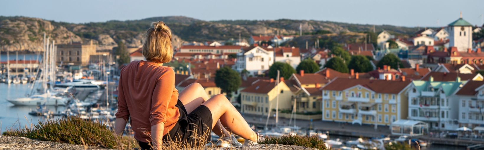 Woman sitting on a cliff at sunset looking out over a coastal town in Sweden