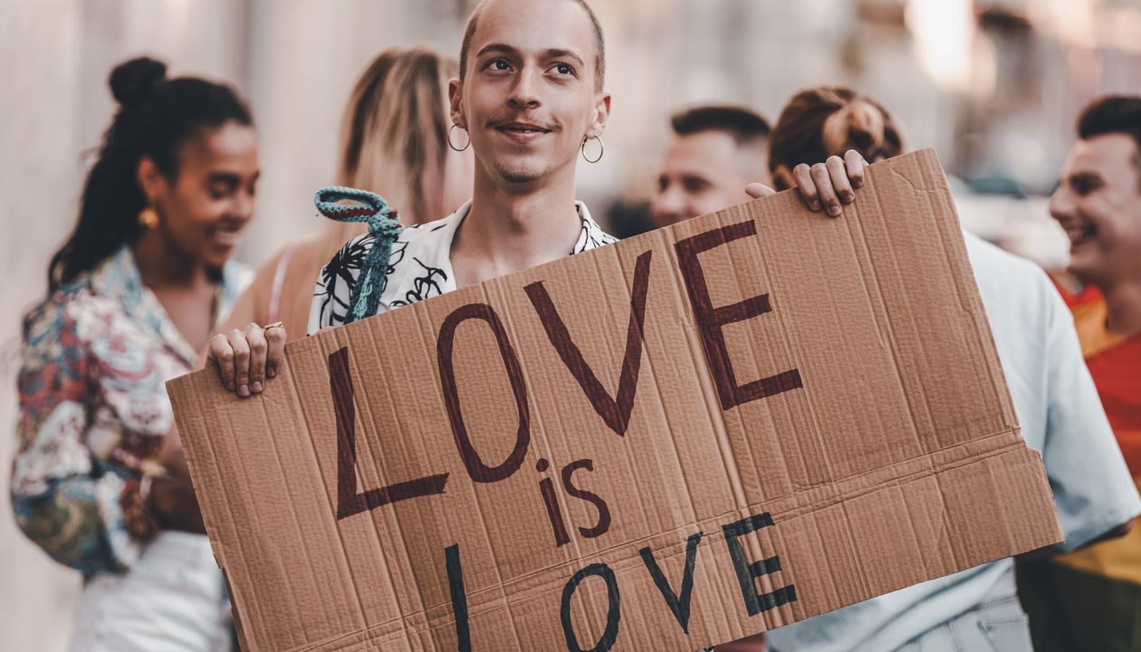 Vyras miesto gatvėje šypsosi ir rodo plakatą „Pride“ eitynėse mieste.