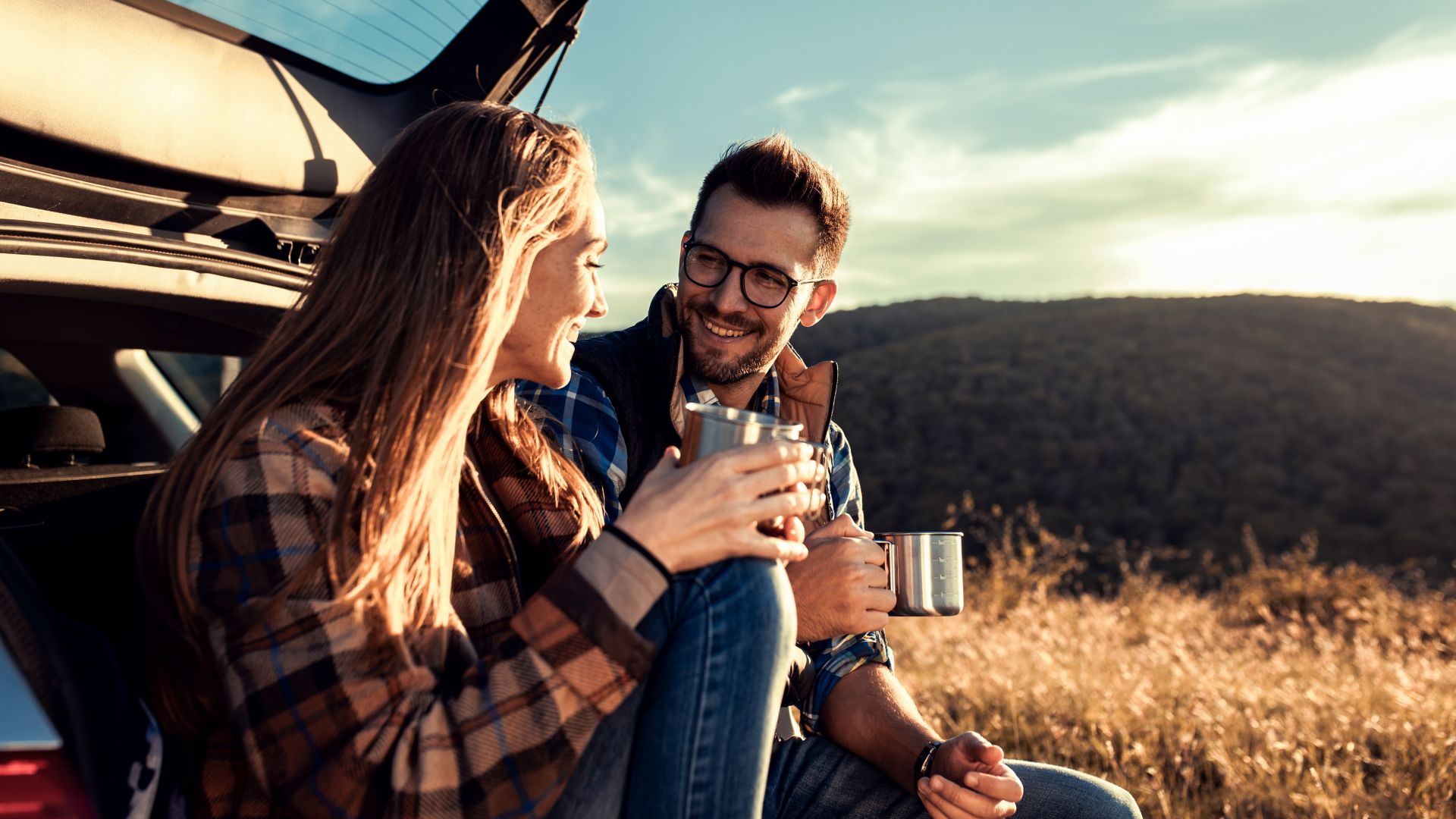 Couple on road trip sitting in trunk of a car resting and drinking coffee.