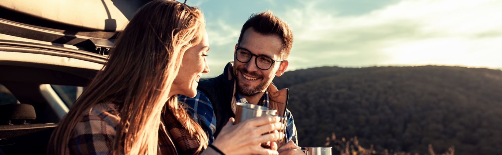 Couple on road trip sitting in trunk of a car resting and drinking coffee.