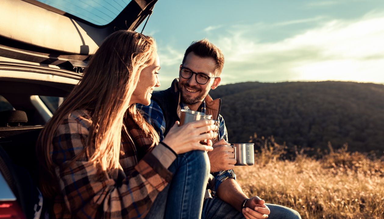 Couple on road trip sitting in trunk of a car resting and drinking coffee.