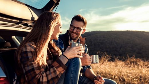 Couple on road trip sitting in trunk of a car resting and drinking coffee.