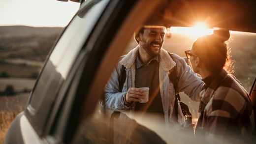 Couple travelling, talking, drinking a coffee watching the sunset from their car. 