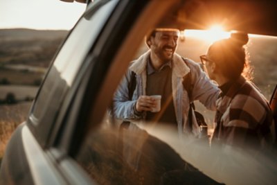 Couple travelling, talking, drinking a coffee watching the sunset from their car. 