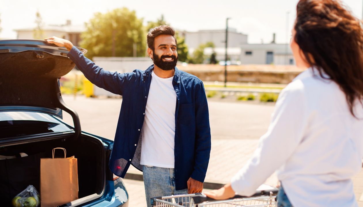 Happy arab couple with shopping cart full of fresh food, packing products into the car on the outdoor parking, woman standing near shopping cart