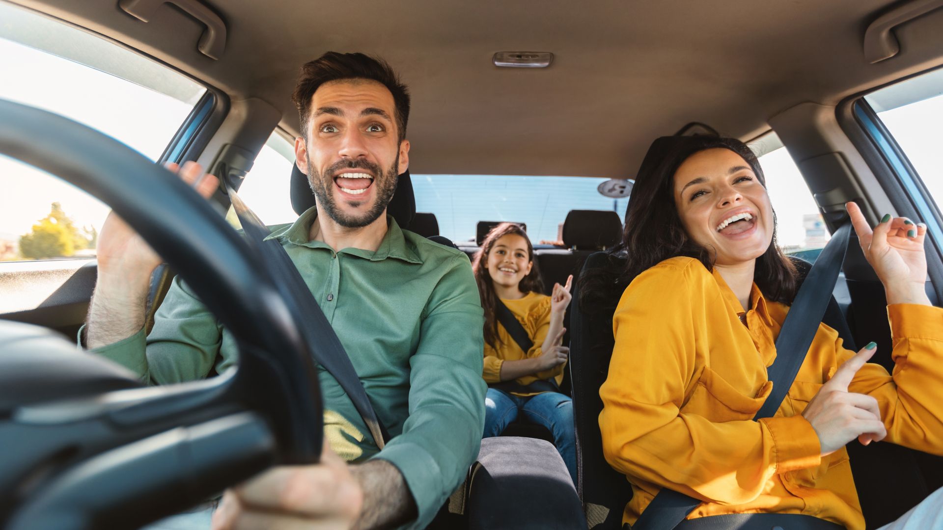 A happy family of dad, mum and young teenage daughter sit, with seatbelts on, in a moving car all singing along to music. 