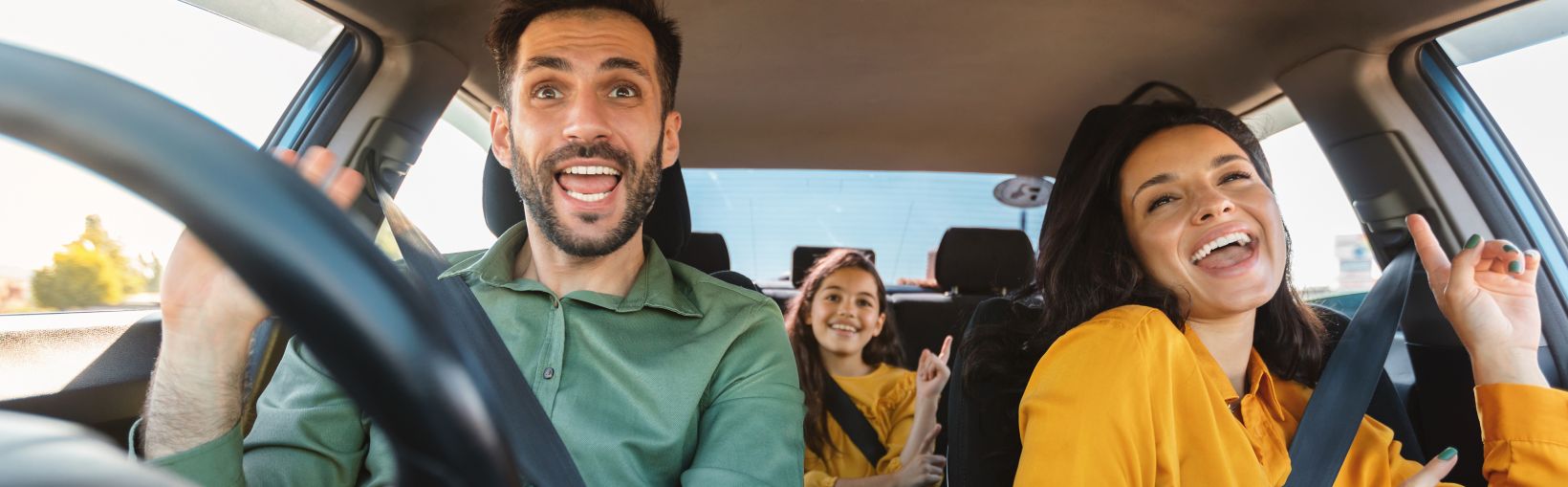 A happy family of dad, mum and young teenage daughter sit, with seatbelts on, in a moving car all singing along to music. 