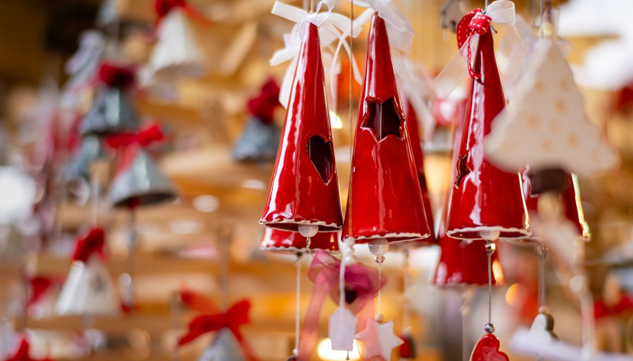 Close-up of glossy red ceramic cone-shaped ornaments hanging in a festive display. The decorations feature cut-out star and heart shapes and small dangling charms. Soft-focus ornaments in the background create a warm, cozy holiday atmosphere. The scene suggests a Christmas market or seasonal craft stall with handmade decorations.