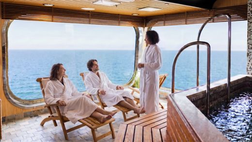 AI-modified image. Three adults in white bathrobes relax in a spa overlooking the ocean through a large panoramic window. Two people sit on wooden lounge chairs while another stands nearby, suggesting a calm conversation. The interior features warm wood panelling, tiled flooring, and a small pool or hot tub area. Soft natural light and the expansive sea view create a tranquil, luxurious atmosphere.