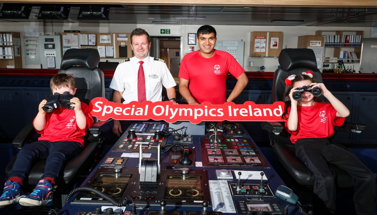 A group of people, including children and adults, are gathered in a ship's control room. Two children in red shirts are seated at the control panel, one holding binoculars, while two adults stand behind them. The group is holding a sign that reads 'Special Olympics Ireland,' and the setting features various control instruments and equipment. The environment appears organized and professional, with a focus on teamwork and inclusion.