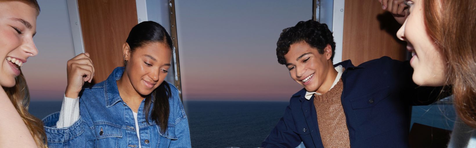 A happy group of 3 young women and 1 young man play a frenetic game of cards at a table in a lounge onboard a Stena Line ferry with the sea and a sunset visible through the tall window beside the table. 