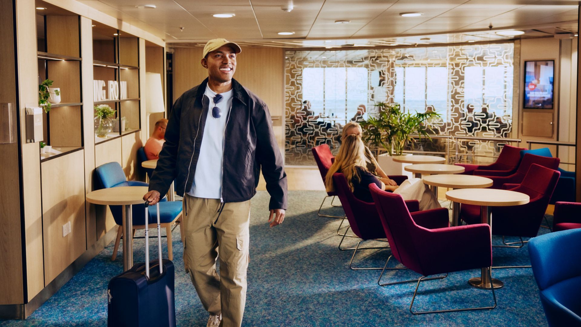 A young man walks through a contemporary lounge pulling a wheeled suitcase. He wears casual travel clothing with a cap, jacket, and sunglasses hanging from his shirt. Other seated passengers relax on colorful chairs and sofas, with tables and indoor plants creating a comfortable atmosphere. Large windows and decorative wall panels suggest a bright, spacious interior on a ship or transport hub.