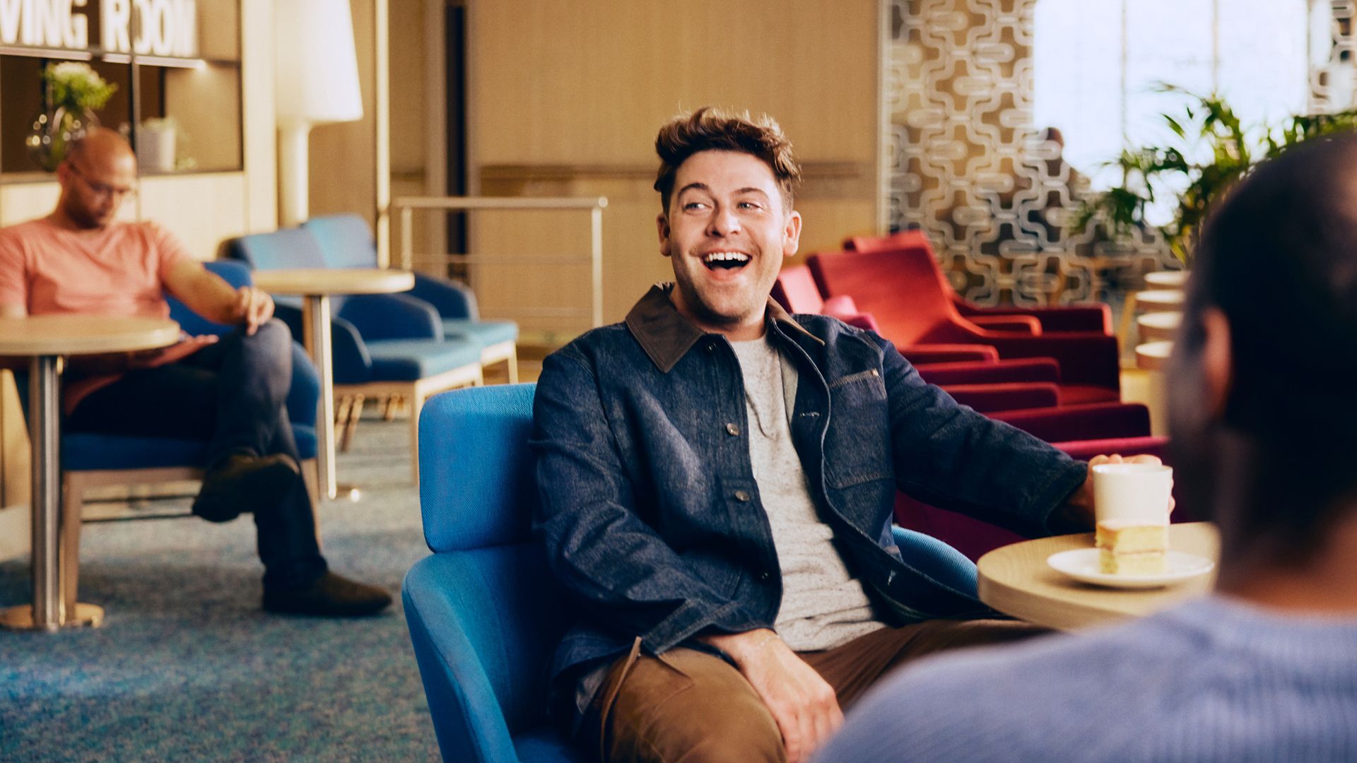 A cheerful man sits in a blue armchair, smiling and talking to someone in a contemporary lounge area. He holds a takeaway coffee cup on the table in front of him, suggesting a casual social moment. The setting features colorful chairs, soft lighting, and a relaxed atmosphere, with other people seated in the background. The overall scene conveys comfort, conversation, and leisure in a stylish public space.