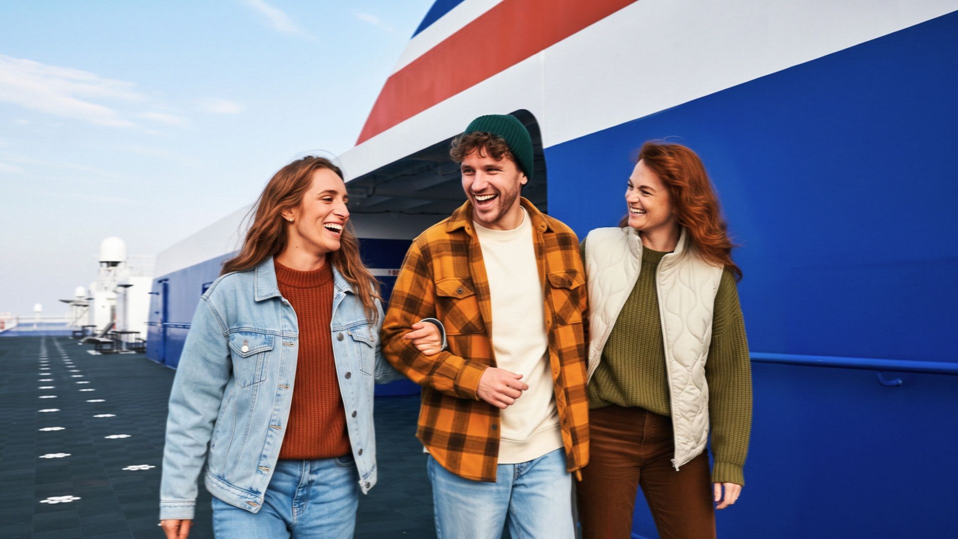 Three young adults walk arm in arm along an outdoor ship deck, smiling and laughing together. The setting appears to be a large vessel with a blue and white exterior and a red stripe, under a clear sky. They wear casual autumn clothing including denim, plaid flannel, knit sweater, and quilted vest, suggesting cool weather. The image conveys friendship, leisure travel, and a relaxed vacation atmosphere.
