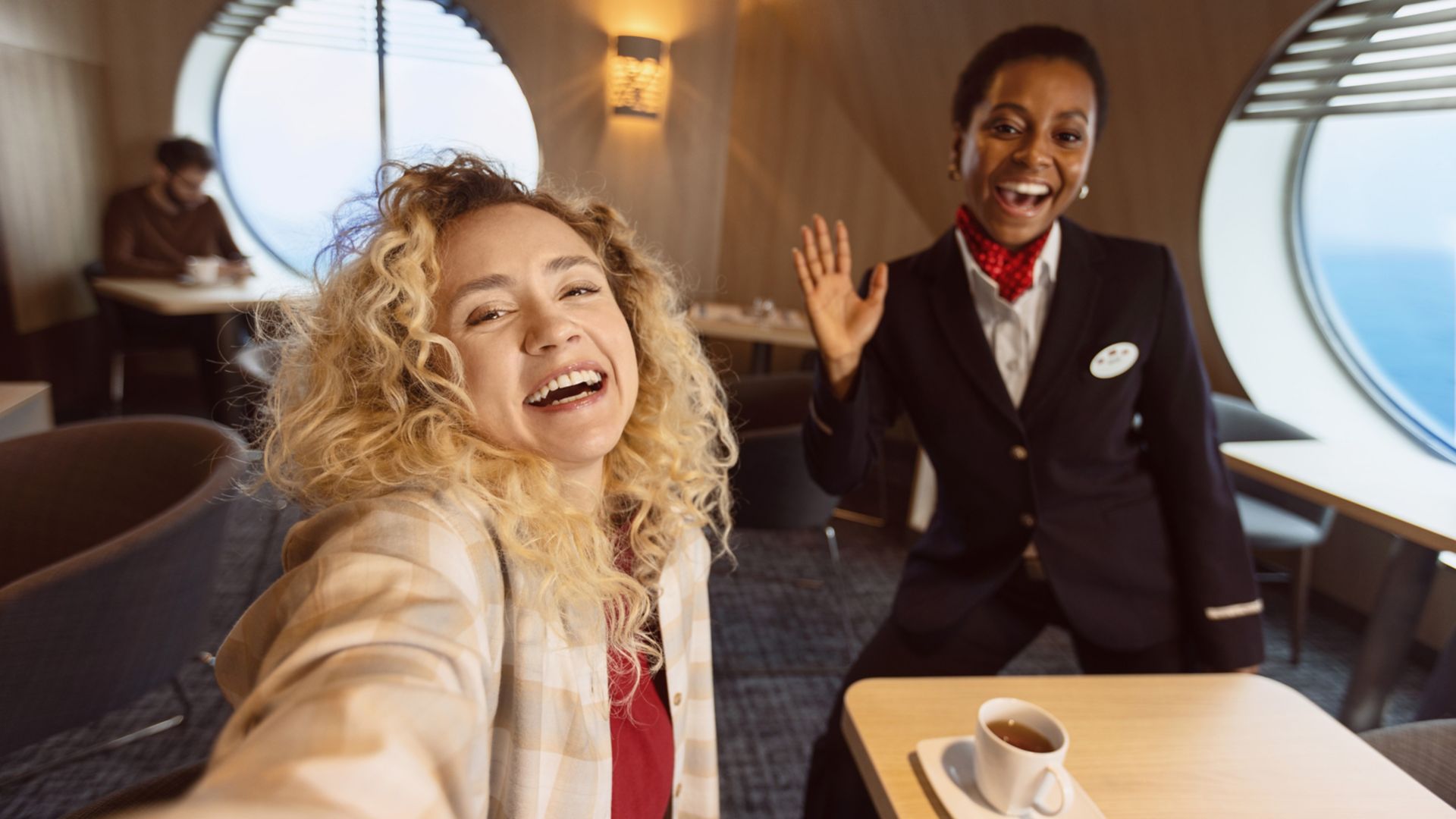 A smiling traveler with curly blonde hair takes a close selfie in a modern lounge area. Beside her, a uniformed crew member with a name badge waves cheerfully toward the camera. The setting appears to be a bright interior with large round windows and a view of the sea outside. Warm lighting, casual seating, and a cup of coffee on the table create a welcoming, relaxed atmosphere.