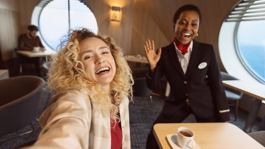 A smiling traveler with curly blonde hair takes a close selfie in a modern lounge area. Beside her, a uniformed crew member with a name badge waves cheerfully toward the camera. The setting appears to be a bright interior with large round windows and a view of the sea outside. Warm lighting, casual seating, and a cup of coffee on the table create a welcoming, relaxed atmosphere.