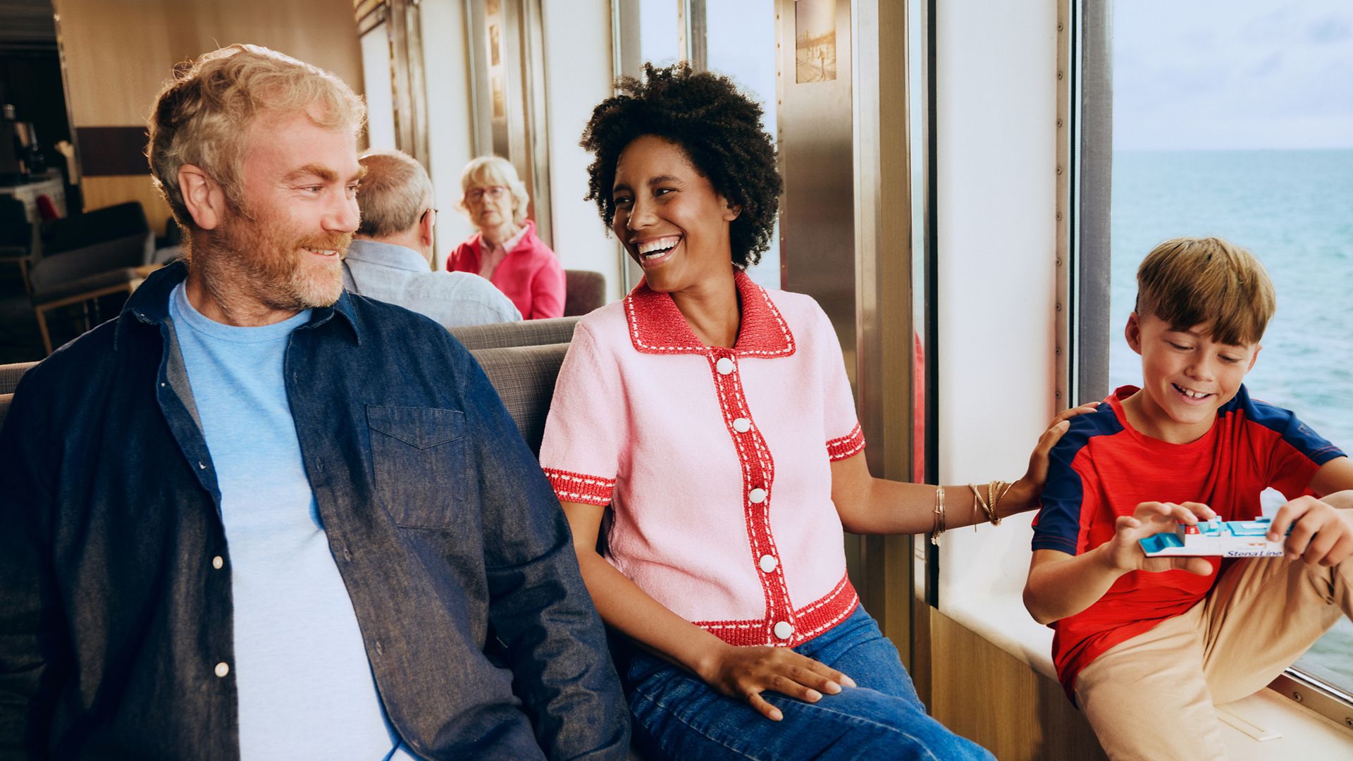 A smiling family sits together on a ferry, enjoying time by the window. The woman in a pink top laughs while interacting with a young boy holding a toy or snack. An older man sits beside them, looking over with a warm expression. Large windows reveal the ocean and bright daylight outside, creating a relaxed travel atmosphere.