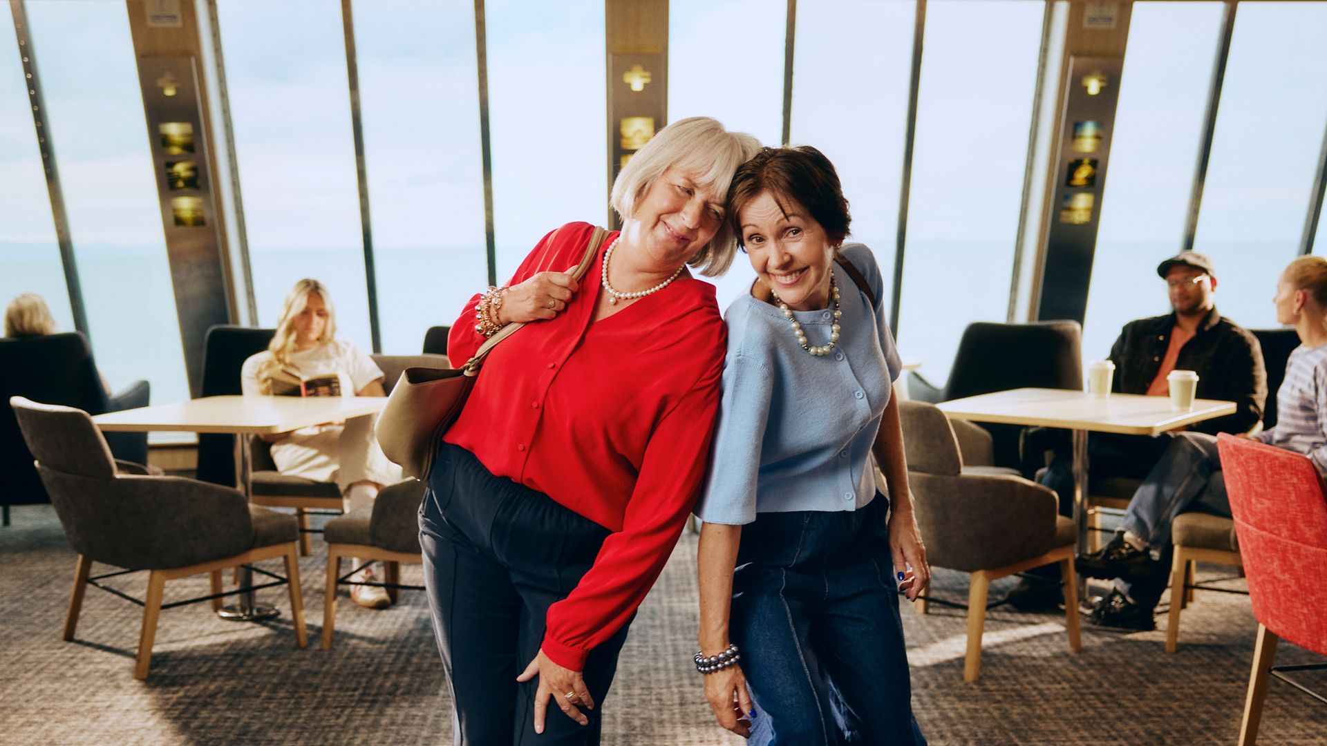 Two older women stand close together and lean playfully toward the camera in a modern café or lounge area. One wears a bright red blouse and the other a light blue top, both smiling warmly and appearing relaxed. In the background, several people sit at tables and chairs near large windows with a view of the sky and water. The scene feels casual and friendly, with soft natural light and comfortable seating creating an inviting atmosphere.