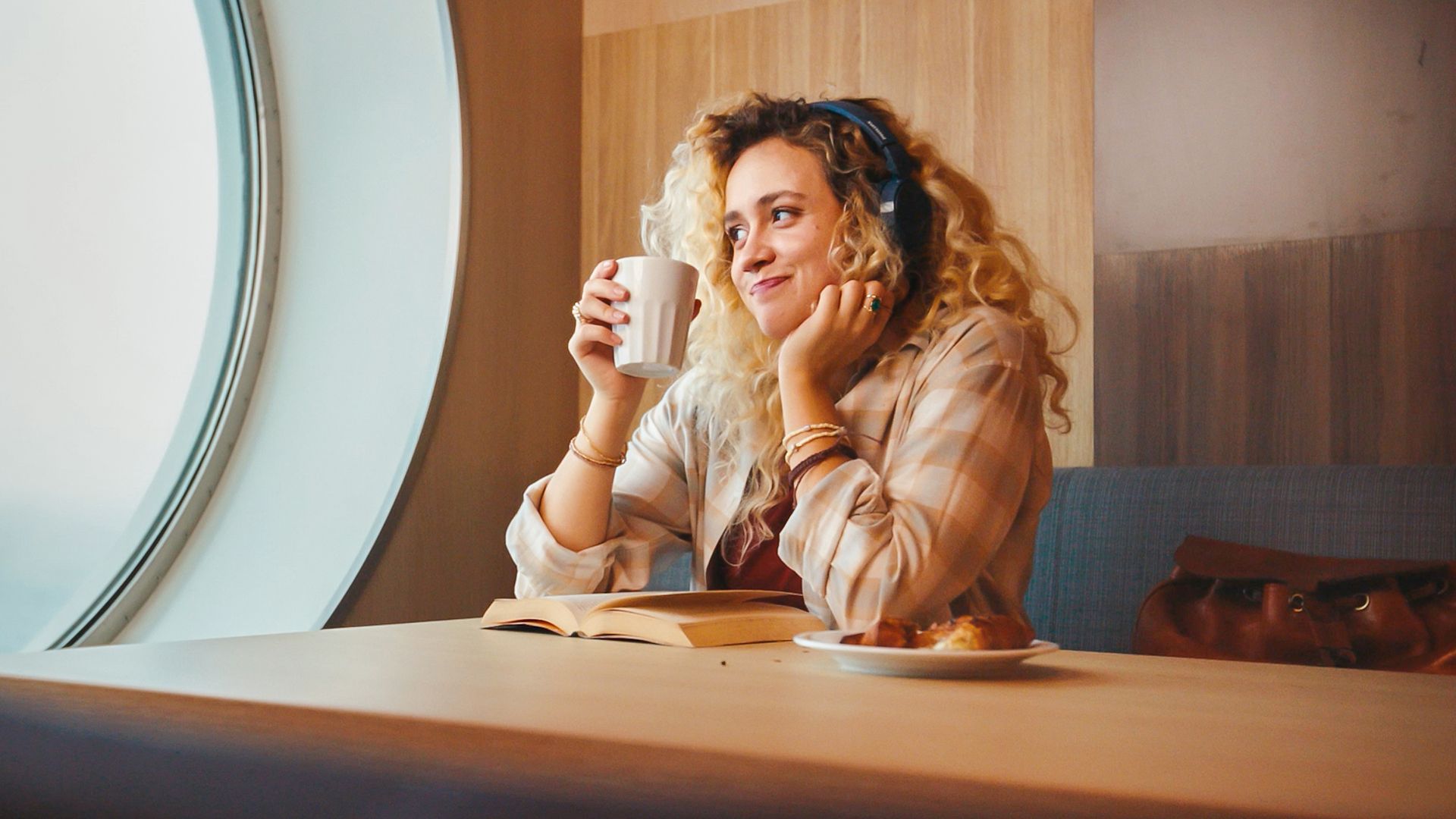 A young woman with curly hair sits at a table by a large round window, holding a white mug and gazing thoughtfully into the distance. She wears headphones and casual clothing, suggesting a relaxed, introspective moment. An open book lies on the table in front of her, along with a small plate of food, hinting at a quiet break or study session. Warm interior lighting and wood paneling create a cozy, comfortable atmosphere.
