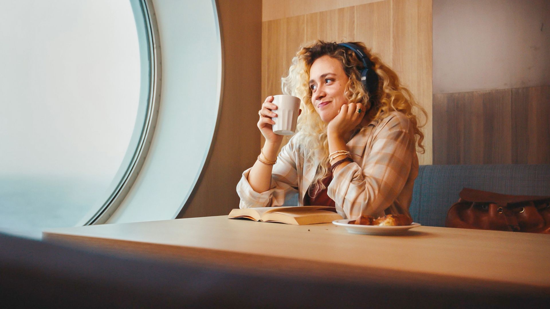 A young woman sits at a table holding a white mug, gazing thoughtfully out of a large round porthole-style window. She appears relaxed and contemplative, with an open book and a pastry in front of her. Warm indoor lighting and wood paneling create a cozy atmosphere, suggesting a quiet moment of leisure or travel. The scene emphasizes comfort, daydreaming, and peaceful solitude.