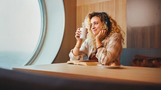 A young woman sits at a table holding a white mug, gazing thoughtfully out of a large round porthole-style window. She appears relaxed and contemplative, with an open book and a pastry in front of her.