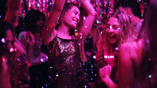 Two young women dance and laugh together at a lively party. They are surrounded by shimmering confetti and metallic streamers under vibrant pink and purple lights. One woman wears a sparkling sequin dress, while the other cheers with her hands raised. The