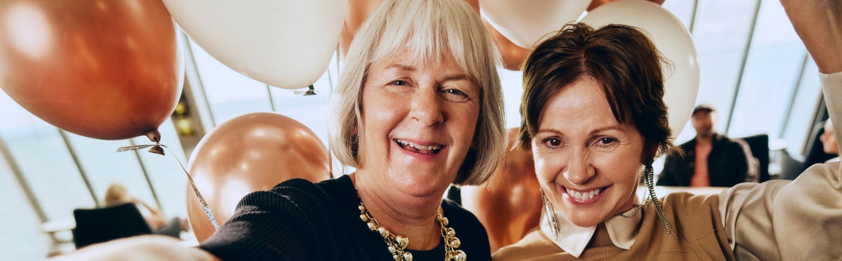 Two smiling women pose closely together for a selfie at an indoor celebration. They stand in front of large white and copper-colored balloons, suggesting a festive event or party. The older woman wears a dark dress with a statement necklace, while the younger woman is dressed in a tan outfit. Warm lighting and soft focus in the background create a cheerful, intimate atmosphere.