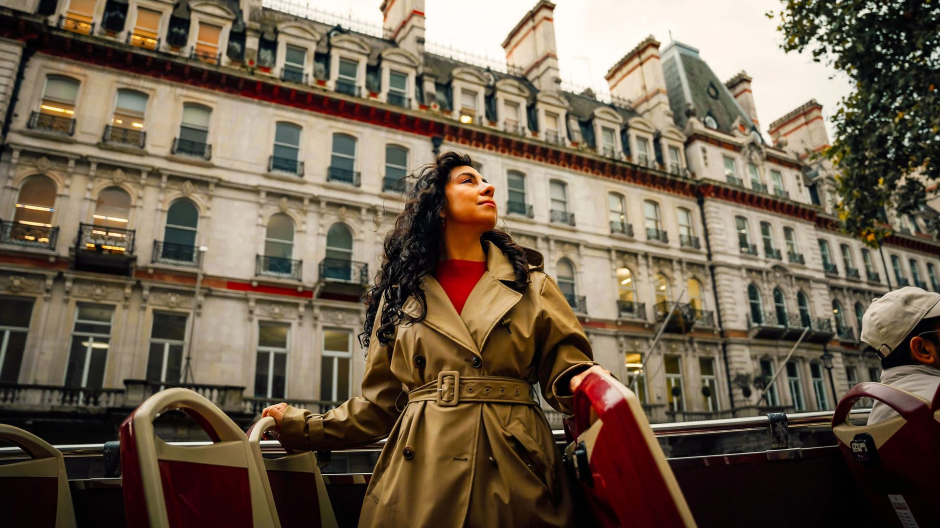 Femme en trench beige profitant d’une visite touristique en bus panoramique, avec une architecture historique en arrière-plan.