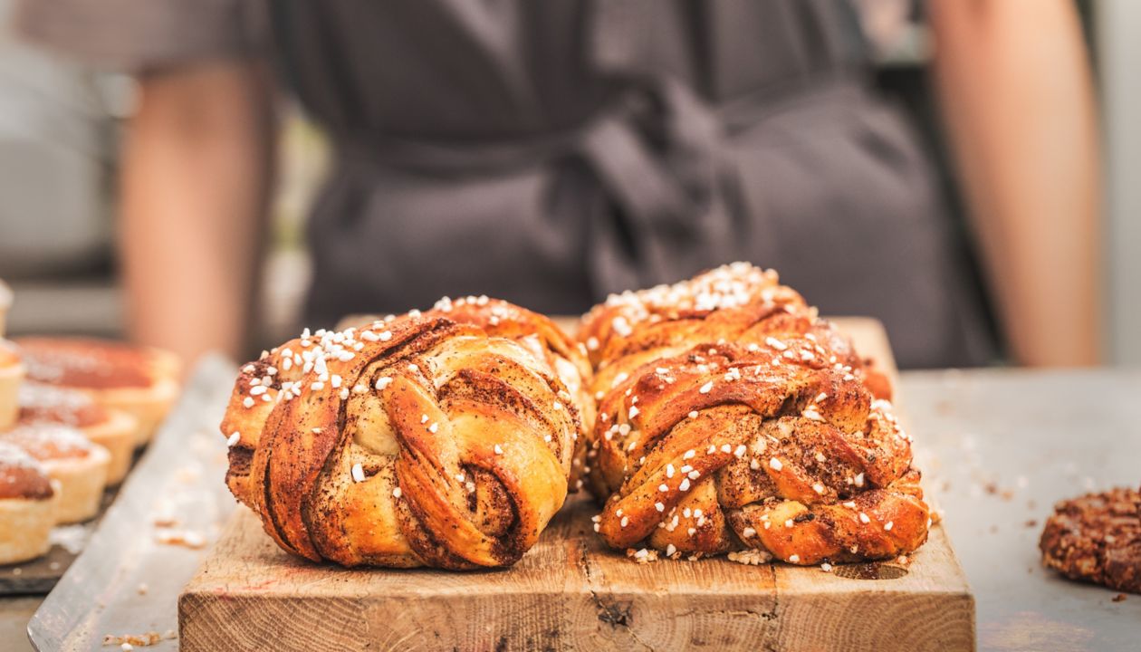 Twisted traditional Swedish cinnamon rolls  at a café. The sweet buns are on a wooden chopping board, and there is an  unrecognizable person wearing an apron in the background.