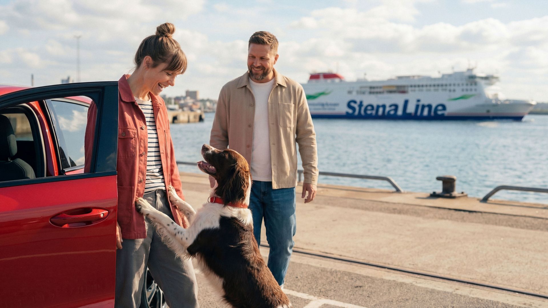 A couple stands beside a red car at a harbour while their dog jumps up playfully. The scene overlooks calm water with a large Stena Line ferry in the background. The people appear relaxed, dressed in casual jackets and jeans on a bright, partly cloudy day. The image conveys travel, companionship, and anticipation of a ferry journey.