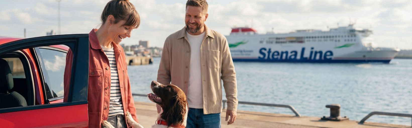 A couple stands beside a red car at a harbour while their dog jumps up playfully. The scene overlooks calm water with a large Stena Line ferry in the background. The people appear relaxed, dressed in casual jackets and jeans on a bright, partly cloudy day. The image conveys travel, companionship, and anticipation of a ferry journey.