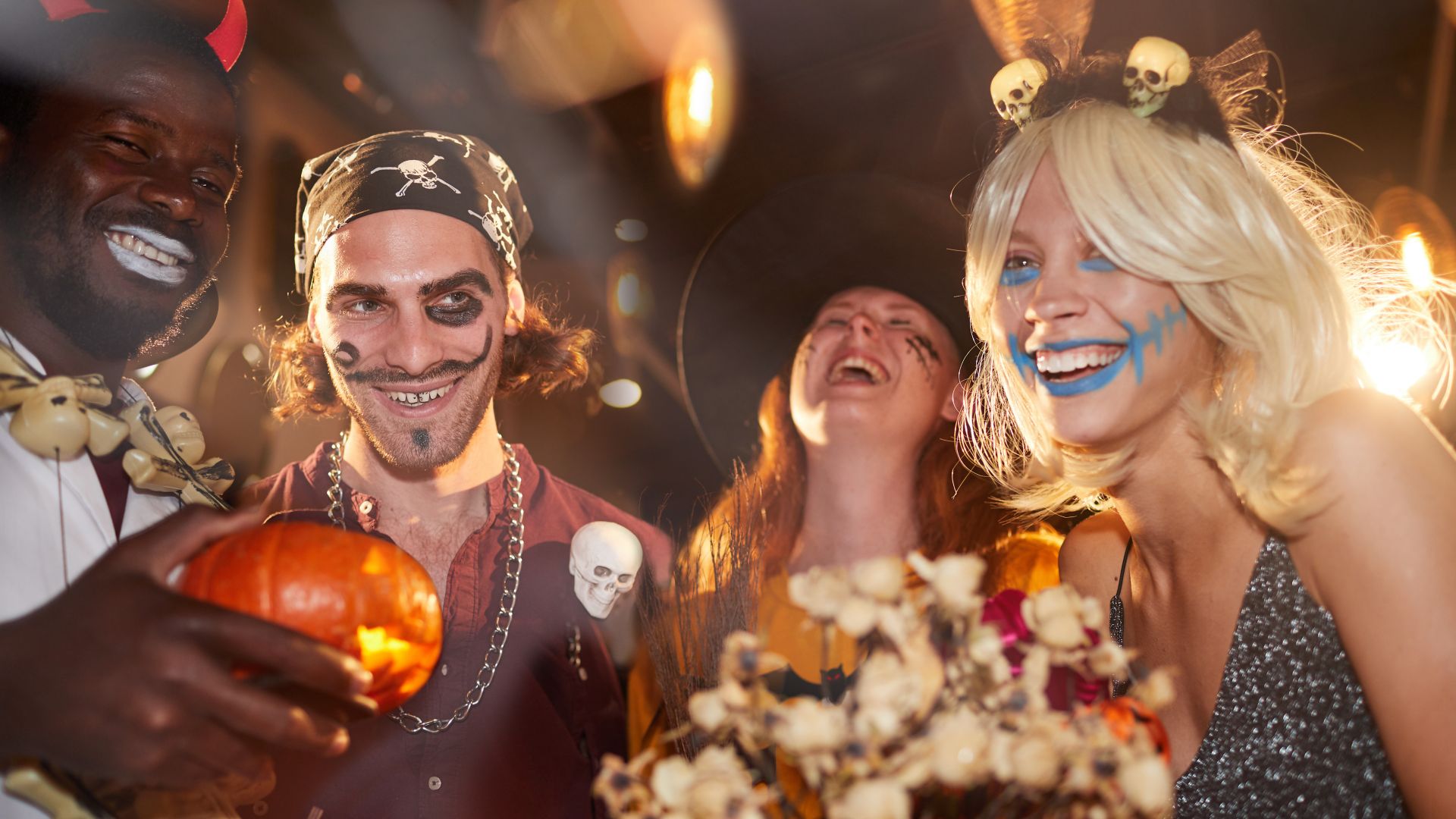 Grupa przyjaciół w halloweenowych kostiumach śmieje się razem na imprezie, trzymając dynię i kwiaty, świętując jesień.