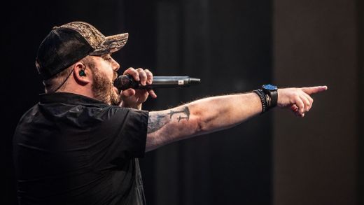 Noel Boland of The Luke Combs Experience dressed in black t-shirt and a black baseball cap sings onstage whilst pointing to the crowd.