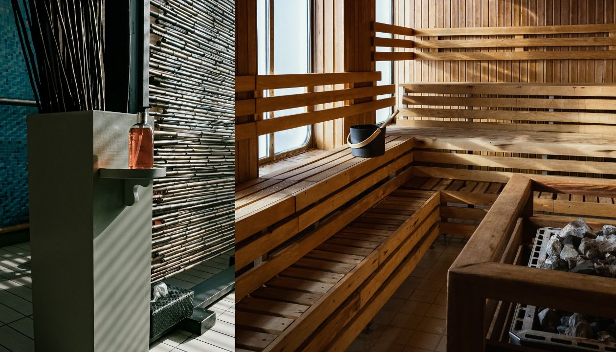 A contemporary sauna interior features tiered wooden benches and a metal heater filled with stones. Sunlight streams through a window, casting warm shadows across the wood paneling. On the left, a tiled area with vertical wooden slats and a minimalist pedestal adds a spa-like touch. A black bucket and ladle rest on one of the benches, emphasizing the relaxation and wellness atmosphere.