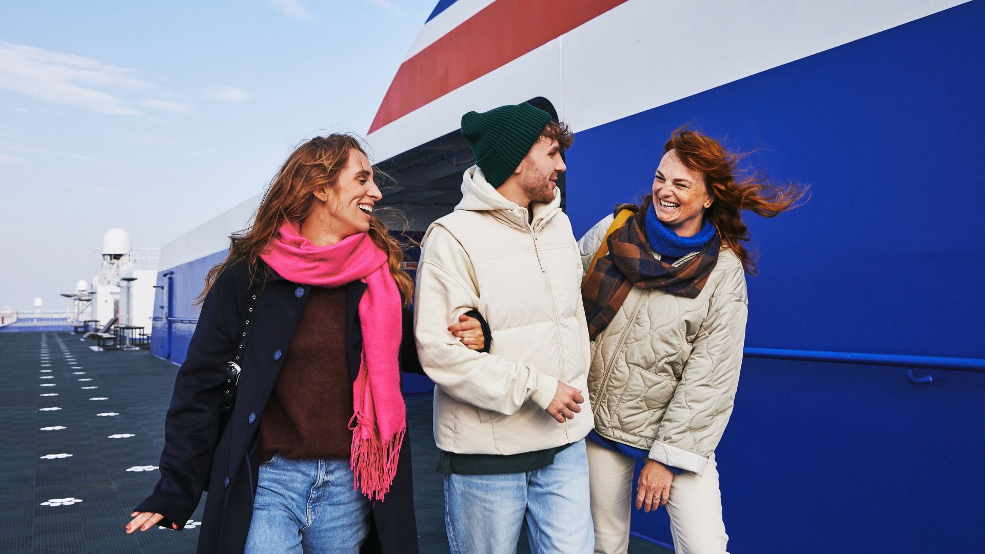 Three people are walking together on the open deck of a large ship or ferry. They are dressed in warm winter clothing, including scarves, jackets, and hats, suggesting a cold or windy day. The background features a bold blue and white ship exterior with a red diagonal stripe. The group appears to be enjoying a casual outing, with relaxed and cheerful body language.