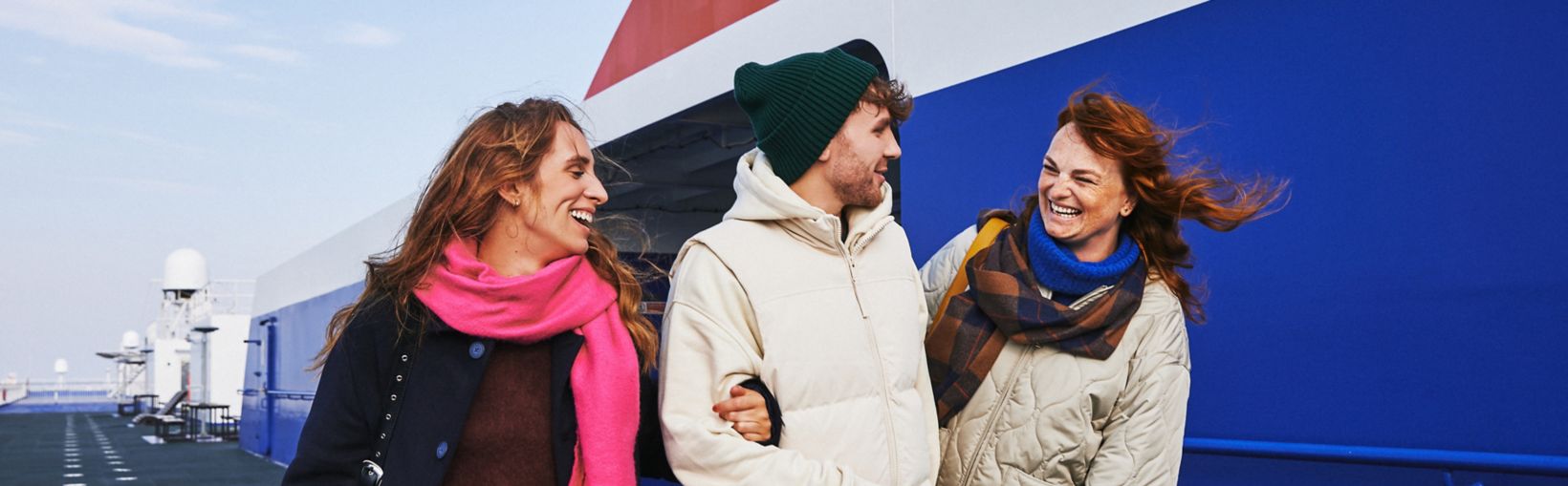 3 friends in warm coats and scarves link arms whilst standing ojn an outside deck of a Stena Line ferry.