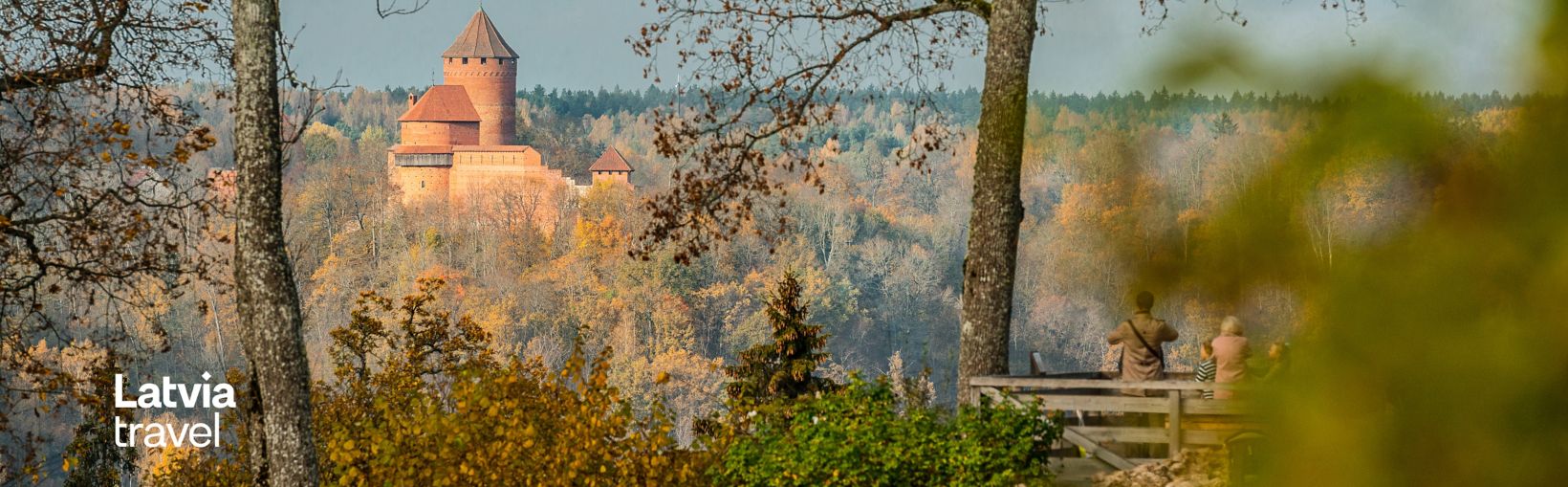 Utsikt över Turaidas slott i nationalparken Gauja i Lettland, omgivet av höstfärgade träd, med besökare som beundrar landskapet från en träplattform.