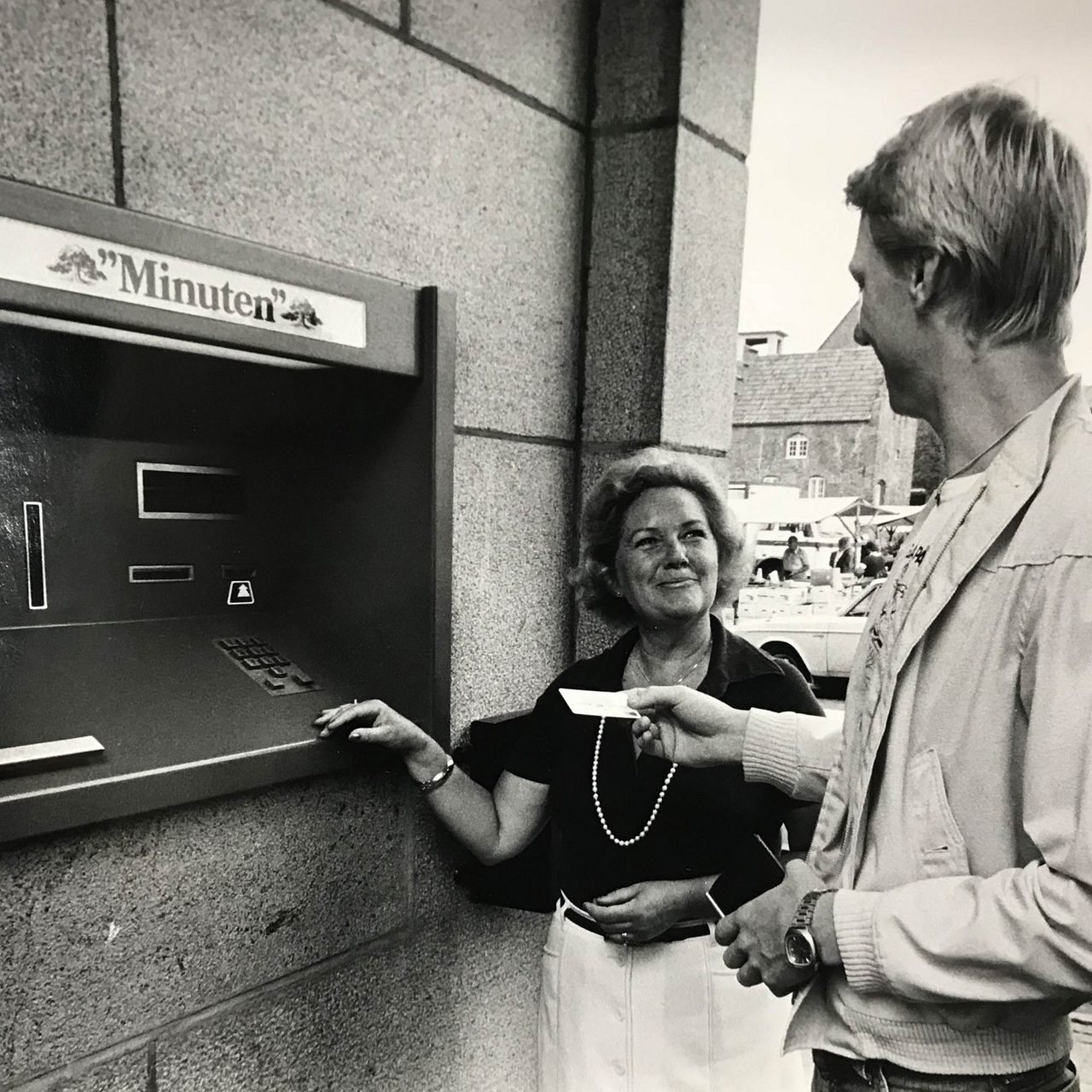 A happy woman and man standing in front of a atm called Minuten. The man is holding a bank card