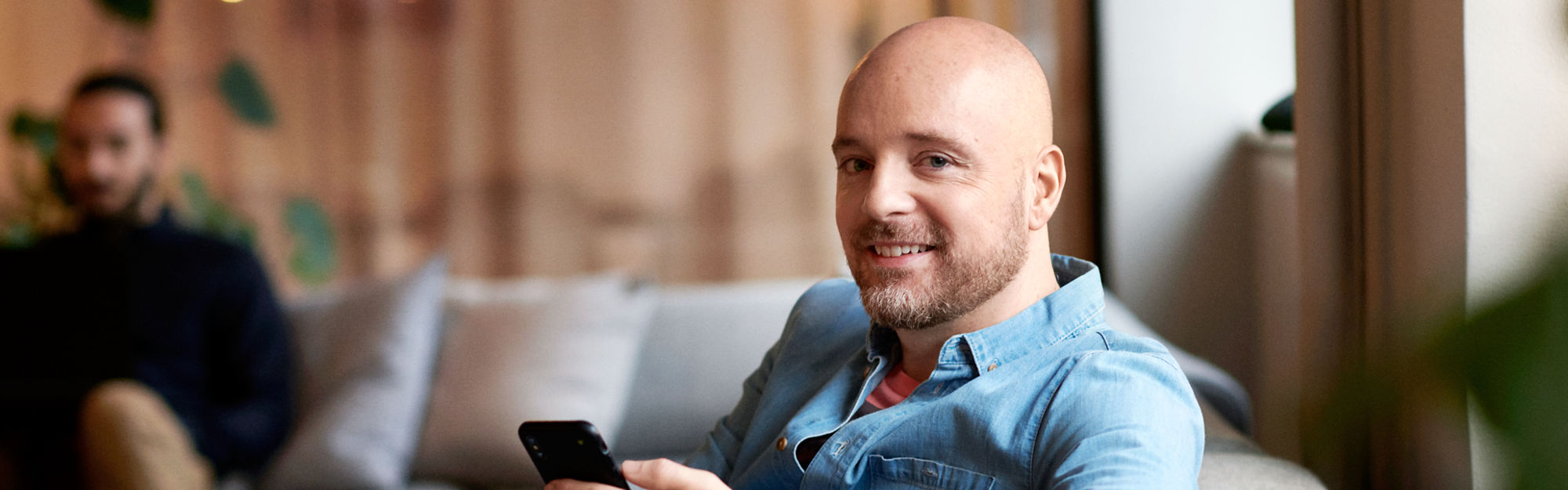 Man in a sofa with a mobile, smiling at the camera.