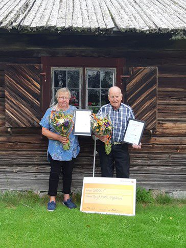 Två äldre personer står utanför ett hus av trä, de håller blommor och diplom i händerna