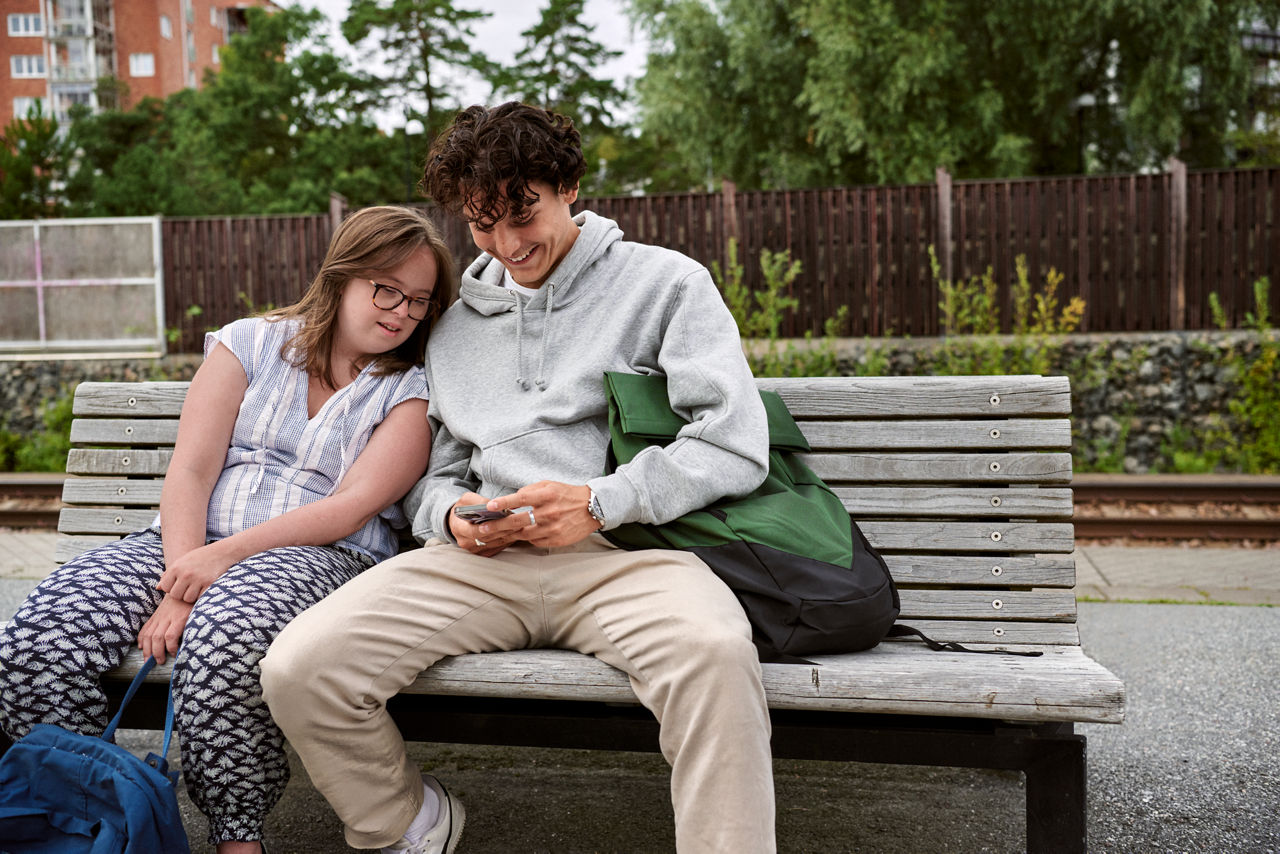 Flicka och tonårspojke på en bänk på en tågstation som tittar på en mobil.
