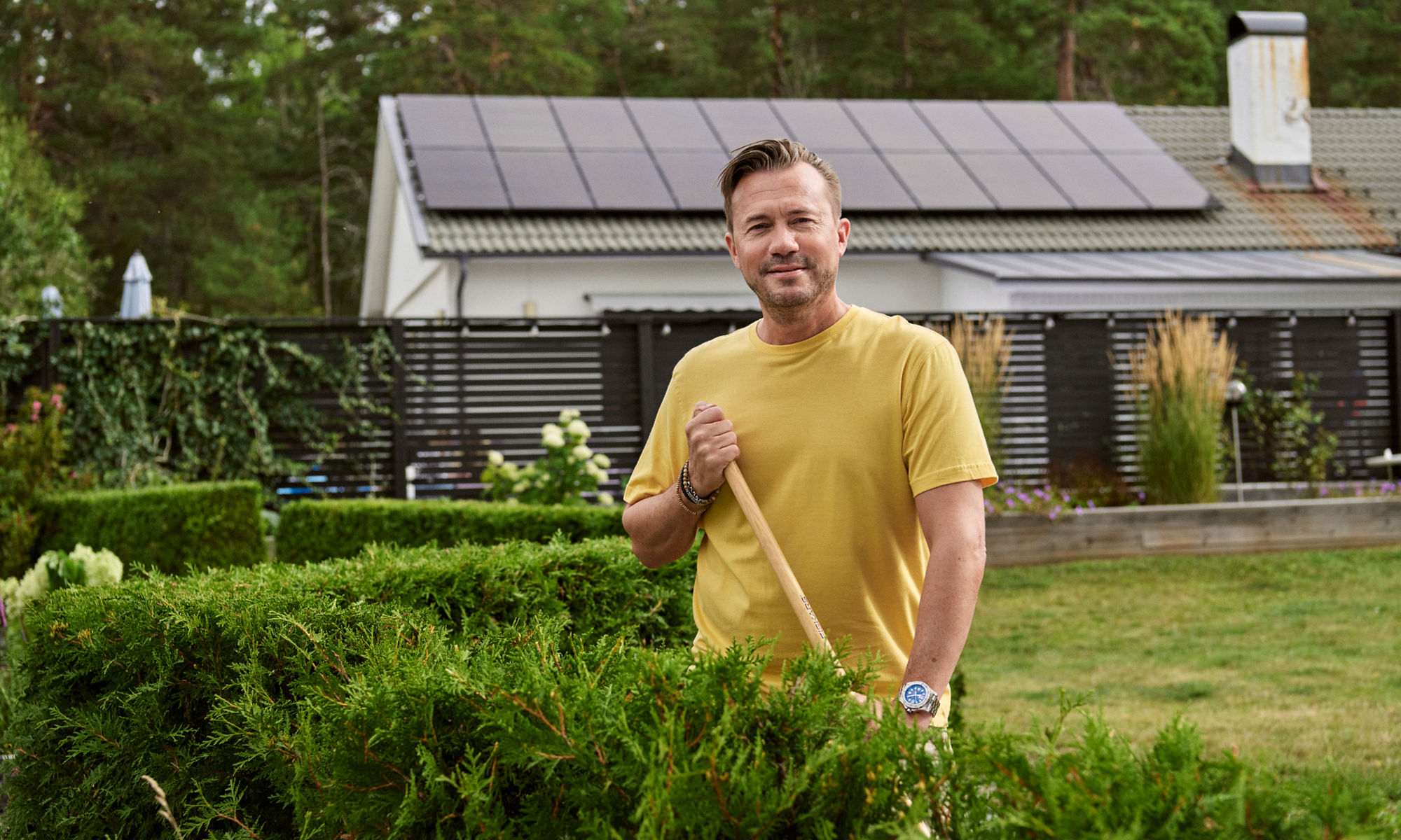 Man klipper häcken i trädgården. Solpaneler syns i bakgrunden.