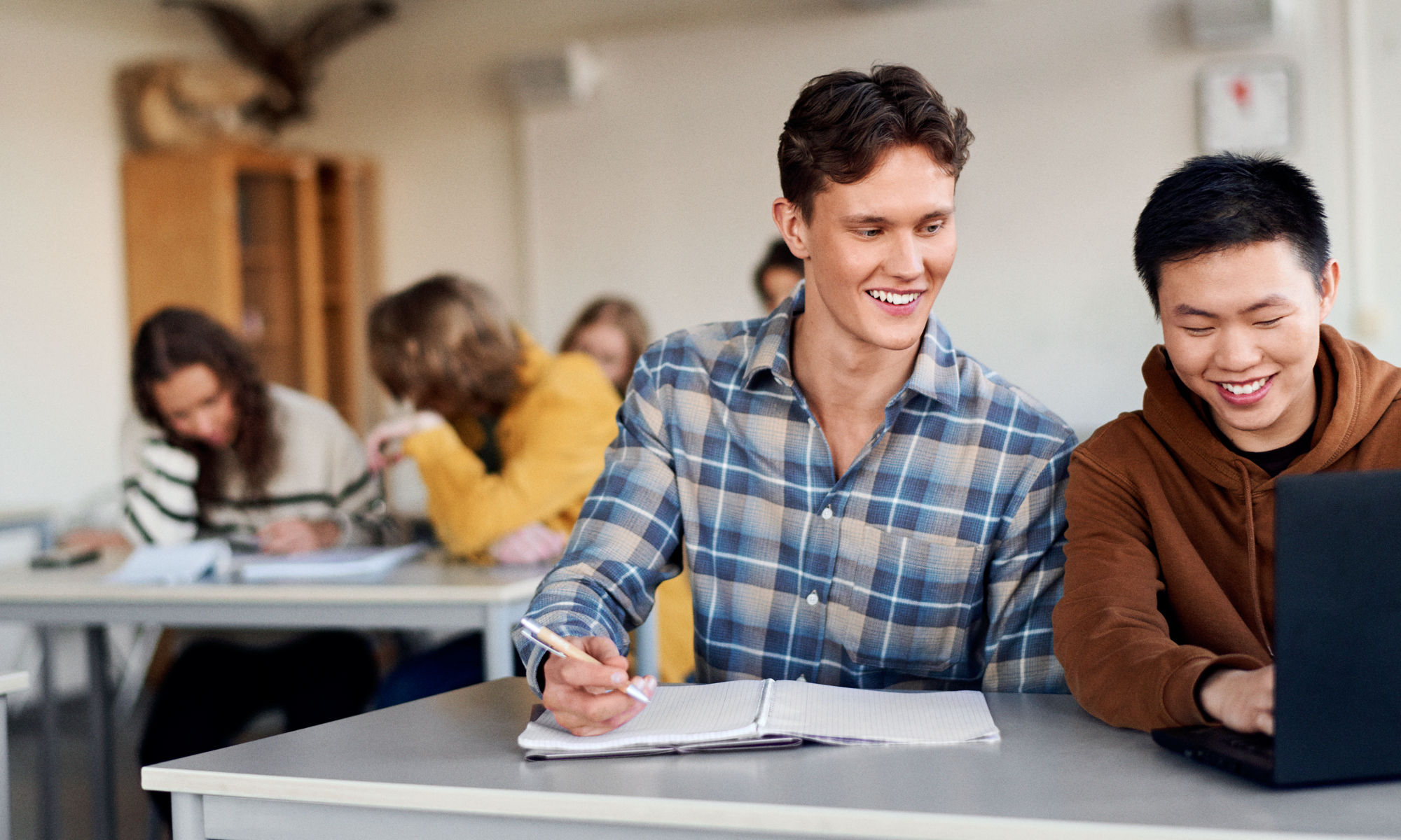 Two male students studying at a desk in classroom, smiling and laughing