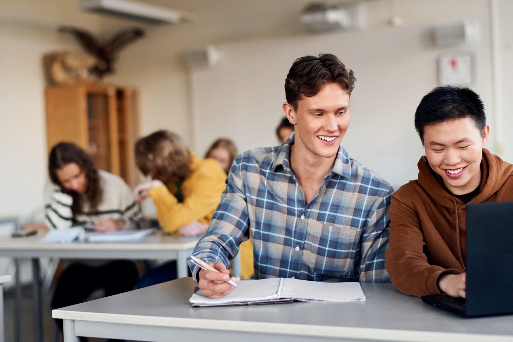 Två manliga studenter pluggar vid ett skrivbord, ler och skrattar